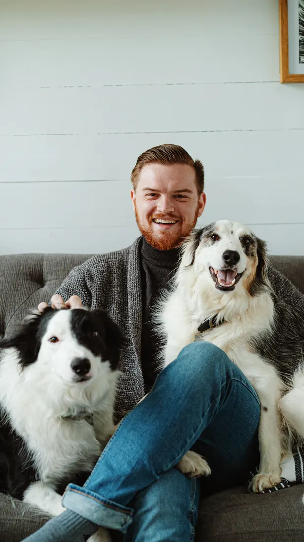 A smiling man sitting on a couch with two happy dogs